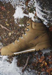 Tan hiking boot with black laces on snowy forest ground with brown leaves, rocks, and a moss-covered tree root.