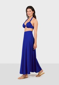 Modré bikiny s riasením a na krku; zodpovedajúca dlhá, flowy sukňa. Materiál je hladký a elastický. Modelka má na sebe zlaté sandále.