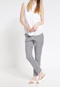 Chemise sans manches blanche à boutons, pantalon à motif vichy noir et blanc, et sandales blanches à enfiler. Tissu lisse, coupe ajustée.