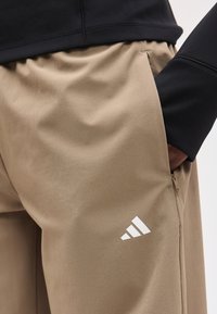 Άτομο που φοράει μπεζ παντελόνια Adidas με μαύρο τοπ, το χέρι στην πλαϊνή τσέπη, ορατό λογότυπο Adidas στα παντελόνια κοντά στην τσέπη με φερμουάρ.