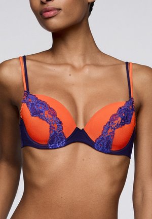 Vrouw die een oranje en paarse kanten bh met dubbele schouderbanden draagt op een lichtgrijze achtergrond.