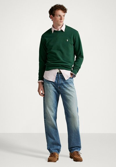 Maglione verde in cotone sopra una camicia rosa chiaro con colletto, abbinati a jeans blu slavati e scarpe di camoscio marroni. Dettaglio semplice del logo sul maglione.