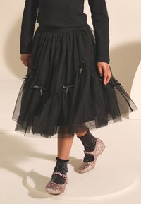 Gonna di tulle nera con volant a strati, arricchita da fiocchi neri. Abbinata a scarpe Mary Jane rosa scintillanti e calzini neri glitterati.