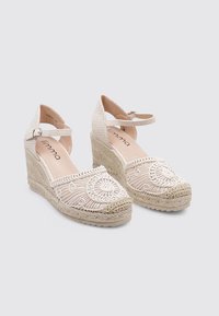 Espadrilles de cuña con una parte superior de tela trenzada en color beige, entresuela de yute trenzado y patrones circulares bordados en la parte frontal. Correa ajustable en el tobillo.
