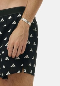 Svarta simshorts med ett upprepande mönster av vita trianglar, med en svart midja. En arm med ett silverarmband är synlig.