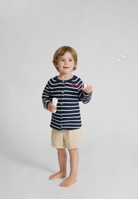 Navy- und weiß gestreifter Strickcardigan mit Holzknöpfen, kombiniert mit beigen Shorts. Das Kind hält einen Seifenblasenstab und zeigt eine Blase.
