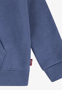 Modra dukserica iz mehkega blaga, z ribastimi manšetami in robom, z stranskim žepom in majhno rdečo oznako na šivu.