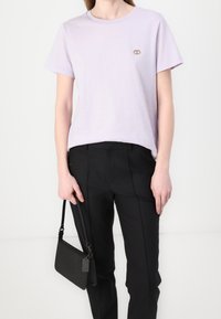 T-shirt en coton lavande avec un col rond et des manches courtes, arborant un petit logo doré. Assorti à un pantalon noir sur mesure et un sac noir.
