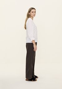 Blouse blanche à manches longues avec des détails brodés, associée à un pantalon large marron et des sandales à enfiler noires en fausse fourrure. Texture lisse.