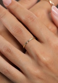 Dünner goldener Bandring mit glatter Oberfläche und einem kleinen Diamantakzent. Hand ruht auf einer strukturierten Oberfläche.