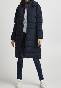 Femme portant un long manteau en duvet bleu marine sur un jean et des baskets blanches, se tenant devant un fond clair.