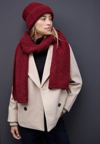 Abrigo beige de doble botonadura, bufanda y gorro de punto rojos. El gorro está enrollado; la bufanda está envuelta de manera suelta. Top negro debajo; jeans oscuros a la vista.