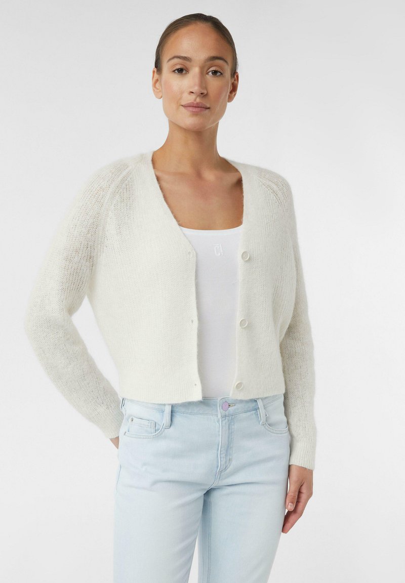 White knitted cardigan with a cropped design, large buttons, and textured sleeves, worn over a white tank top and light blue jeans.