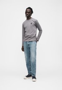 Juodaodis vyras, stovintis su viena ranka kišenėje, dėvintis pilką ilgomis rankovėmis marškinį, šviesiai mėlynas džinsus ir juodus sportinius batelius ant balto fono.