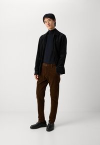 Sort fløjlskjort, marineblå turtleneck, brune fløjlbukser og sorte sko med hardwaredetaljer. Modellen står imod en ensfarvet baggrund.