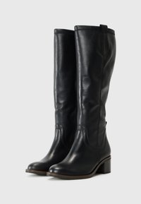 Bottes en cuir noir montantes avec une texture lisse, bout arrondi, talon bas et bloc, et des coutures décoratives sur les côtés.