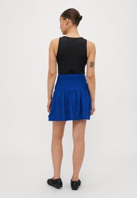 Μαύρο αμάνικο τοπ συνδυασμένο με μίνι φούστα από μπλε δαντέλα, με ρεβάρ ύψους και τιρεδάτη ραφή στο τελείωμα. Μαύρα παπούτσια ολοκληρώνουν το σύνολο.