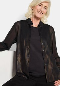 Doorzichtig zwart button-up shirt met een subtiele metallic glans, lange mouwen en klassieke kraag, gelaagd over een effen donkere top.