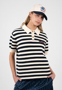 Femme portant un polo en maille à manches courtes rayé crème et noir, une casquette crème avec logo, des boucles d'oreilles anneaux dorées et un pantalon bleu marine, les mains dans les poches.