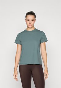 Jovem mulher com o cabelo apanhado em um coque, vestindo uma t-shirt azul-petróleo de manga curta e leggings castanhas, em pé contra um fundo claro e liso.