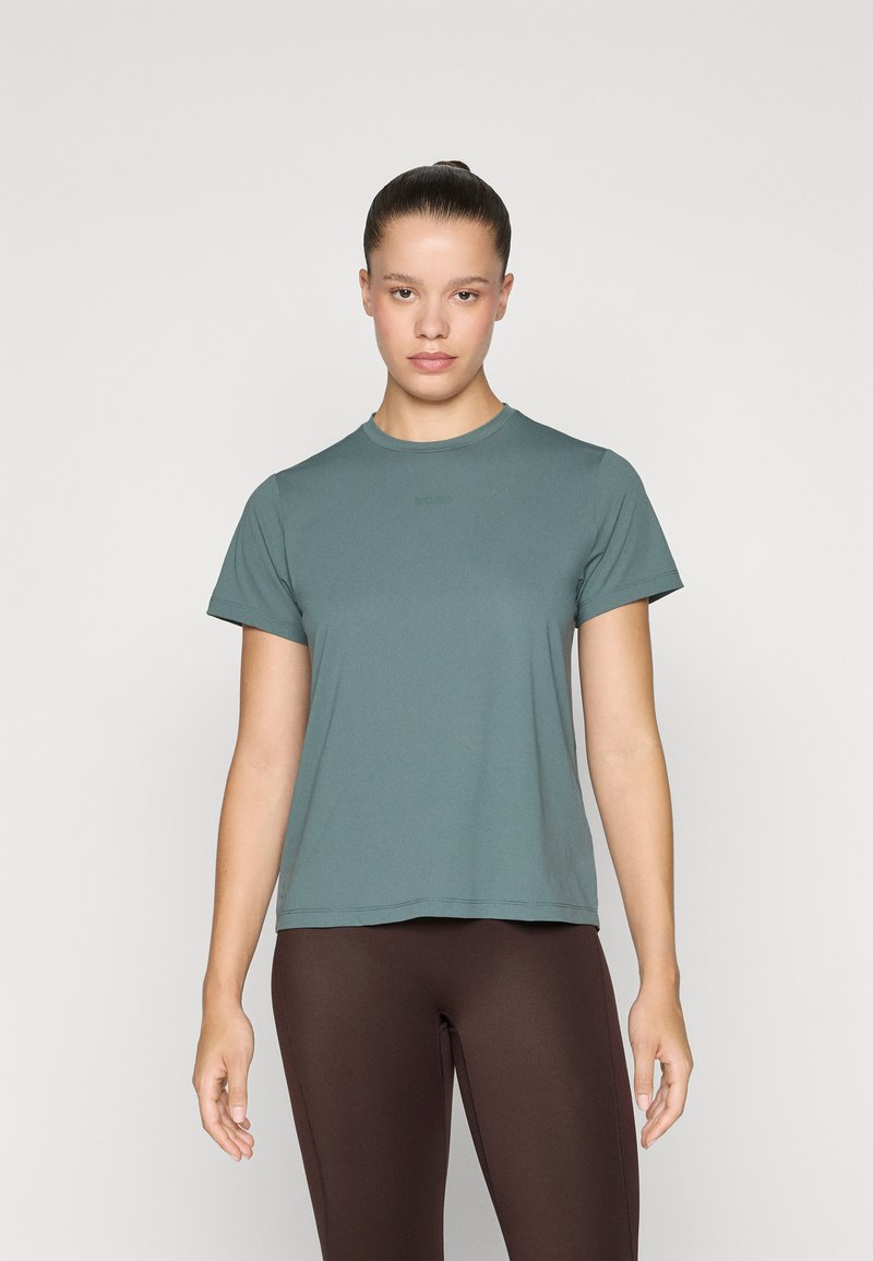 Jovem mulher com o cabelo apanhado em um coque, vestindo uma t-shirt azul-petróleo de manga curta e leggings castanhas, em pé contra um fundo claro e liso.