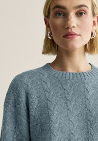 Vaaleansininen neulottu villapaita, jossa kaapelineuloskuvio. Varustettu resorireunuksella kauluksessa ja teksturoidulla kankaalla, jossa on hienovaraisia pilkkuja.