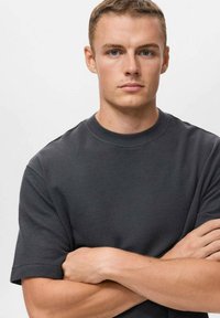 Tmavě šedé bavlněné tričko s krátkými rukávy a kulatým výstřihem, má volný střih a hladkou texturu. Model stojí se zkříženýma rukama.