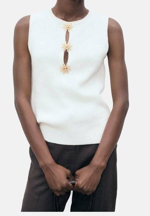 Bílý bezrukávový pletený top s třemi zlatými květinovými akcenty v oblasti výstřihu a malým výřezem. Hladká textura a přiléhavý střih.