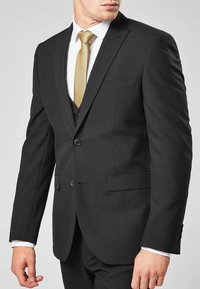 Svart skräddarsydd kostym med en kavaj med två knappar, utskurna slag, en bröstficka och byxor utan veck. En guldfärgad slips kompletterar hela outfiten.