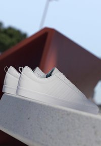 Par de zapatillas blancas limpias de corte bajo colocadas sobre una superficie de piedra clara con una estructura geométrica roja desenfocada en el fondo.