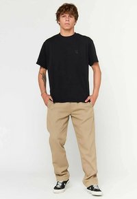 Zwart katoenen T-shirt met een klein logo, gecombineerd met beige broek. De outfit wordt aangevuld met zwart-witte sneakers, staand tegen een neutrale achtergrond.