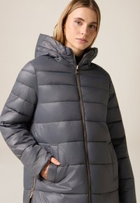 Grå dunjakke med hætte, der har et quiltet design, en frontlynlås og sidelommer. Materialet har en skinnende, glat tekstur.