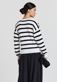 Woman with dark hair in a bun wearing a white sweater with black horizontal stripes and a black skirt, holding a black clutch bag.