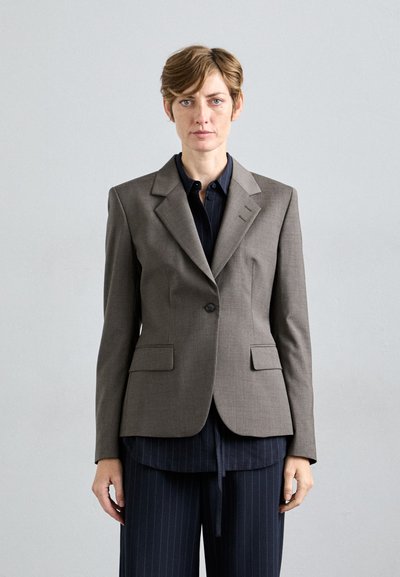 Frau mit kurzer Frisur trägt einen grauen Blazer über einem dunklen Streifenhemd und -hose, steht vor einem schlichten hellgrauen Hintergrund.