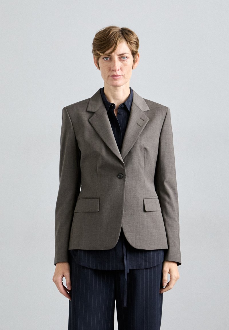 Frau mit kurzer Frisur trägt einen grauen Blazer über einem dunklen Streifenhemd und -hose, steht vor einem schlichten hellgrauen Hintergrund.