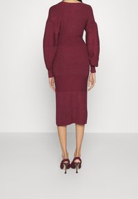 Abito a maglia bordeaux con maniche lunghe e silhouette aderente, caratterizzato da costine testurizzate e lunghezza fino al polpaccio, abbinato a scarpe con tacco alto.