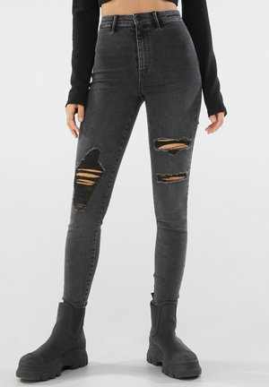 Jean noir slim taille haute avec de larges déchirures usées sur les cuisses, porté avec des bottes noires épaisses et un top court noir.