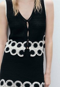 Schwarze Strickweste mit weißen, runden Häkelakzenten am Saum und Knopfverschluss. Verfügt über einen tiefen V-Ausschnitt und ein ärmelloses Design.