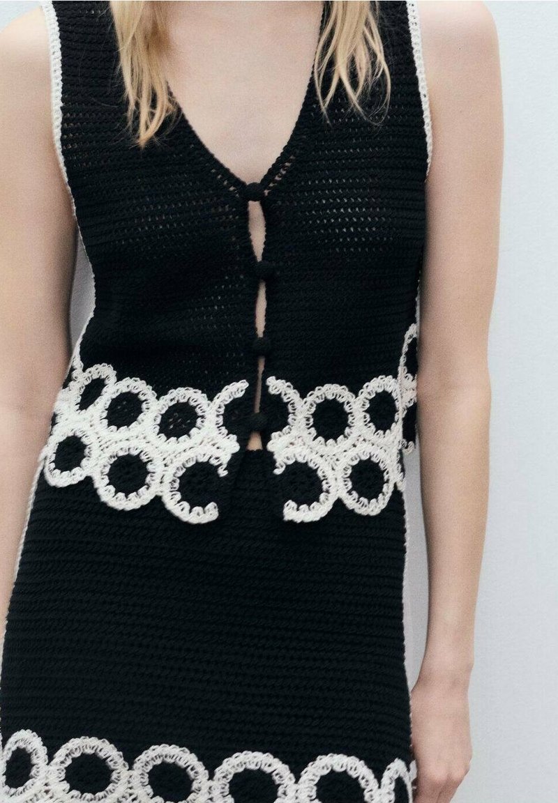 Schwarze Strickweste mit weißen, runden Häkelakzenten am Saum und Knopfverschluss. Verfügt über einen tiefen V-Ausschnitt und ein ärmelloses Design.