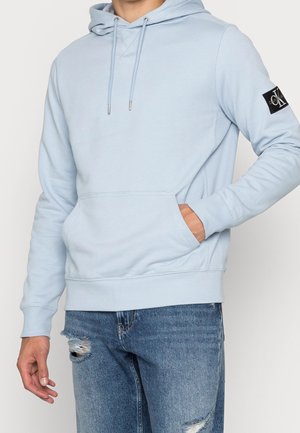 Mężczyzna w jasnoniebieskiej bluzie z kapturem z przednią kieszenią i czarną naszywką z logo na rękawie, w zestawie z podartymi niebieskimi dżinsami.