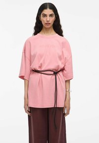 Svetla roza oversized majica s kratkimi rokavi, z napisom "ROUGE EDIT". Slogovno kombinirana z črnim pasom in temno rjavimi širokimi hlačami.