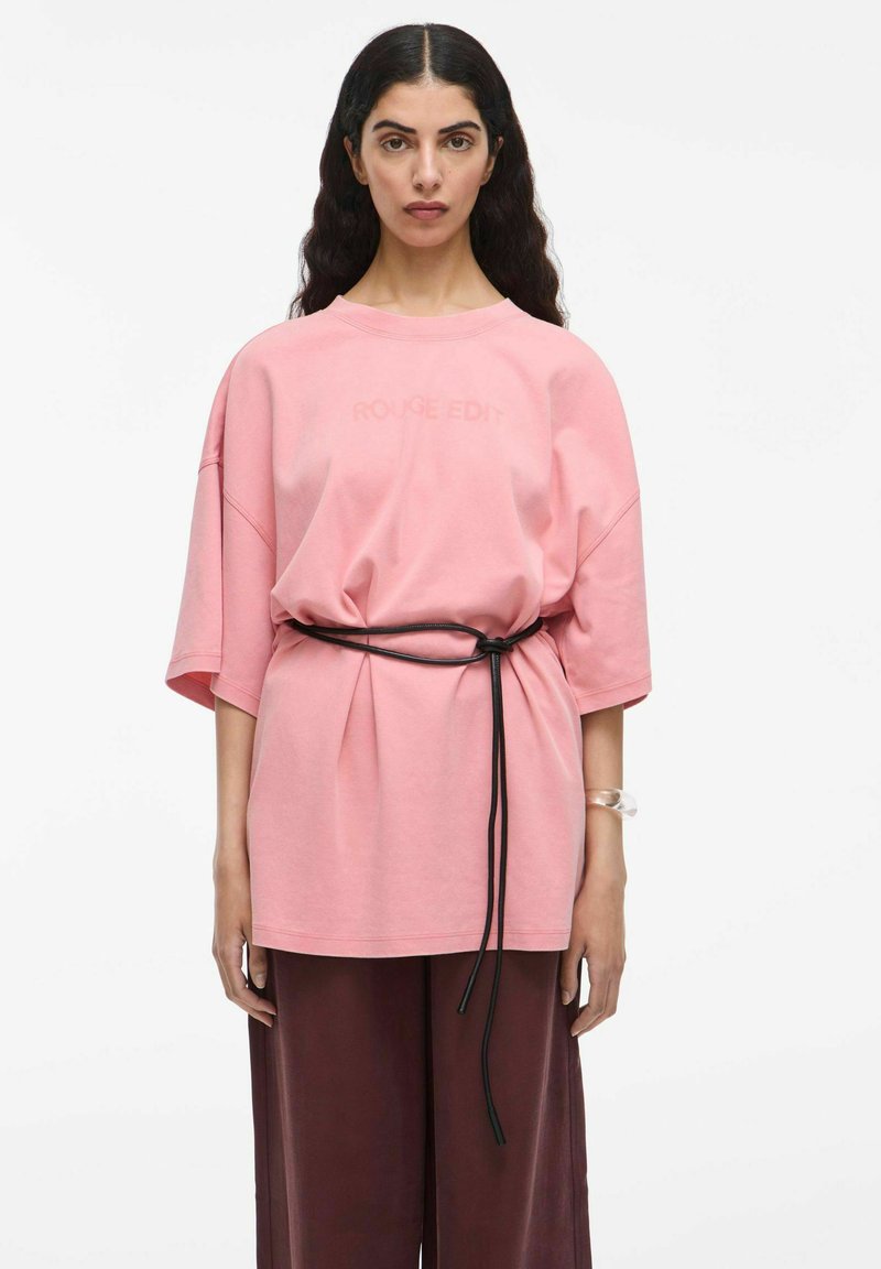 Helles rosa Oversize-T-Shirt mit kurzen Ärmeln, mit "ROUGE EDIT" Druck. Gestylt mit einem schwarzen Gürtel, kombiniert mit dunkelbraunen weit geschnittenen Hosen.