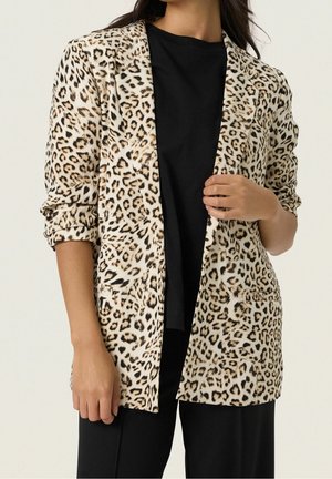 Kvinde iført en lang leopardmønstret blazer over en sort top og sorte bukser, med den ene hånd der justerer blazers front.