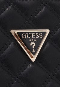 Musta tikattu keinonahkainen käsilaukku, jossa on kultainen kolmionmuotoinen LOGO-KORISTE, jossa lukee "GUESS U.S.A." ja kysymysmerkkikuvio.