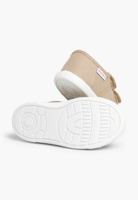 Pantofi dintr-o țesătură bej deschis cu un superior din canva durabil, talpă din cauciuc albă și un sistem de prindere cu arici pentru o îmbrăcare ușoară.