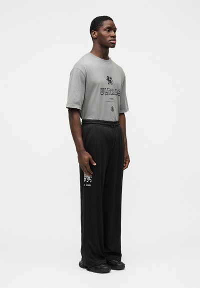 Šedé nadmerné tričko s potlačou "ULTRAS", kombinované s čiernymi voľnými nohavicami s grafickým logom a textom blízko lemu. Čierna obuv.