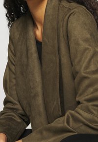 Blazer in un verde oliva, realizzato in tessuto morbido, con colletto sciallato e apertura frontale. Presenta una vestibilità rilassata e una texture liscia, ideale per essere indossato a strati.