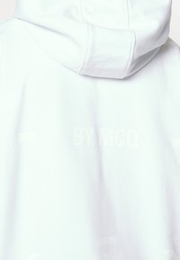 White hoodie in soft fabric features a large hood, with embossed text "BY MCQ" and "LONDON" on the back, showcasing a minimalist design.