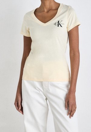 Kobieta ubrana w jasnobeżowy T-shirt Calvin Klein Jeans z dekoltem w serek i białe spodnie, stojąca na jednolitym białym tle.