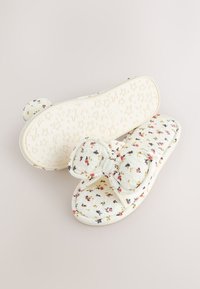 Chaussons doux blancs avec motif floral, nœud rembourré sur le dessus et semelle à motifs étoiles et lune, conçus pour le confort intérieur.